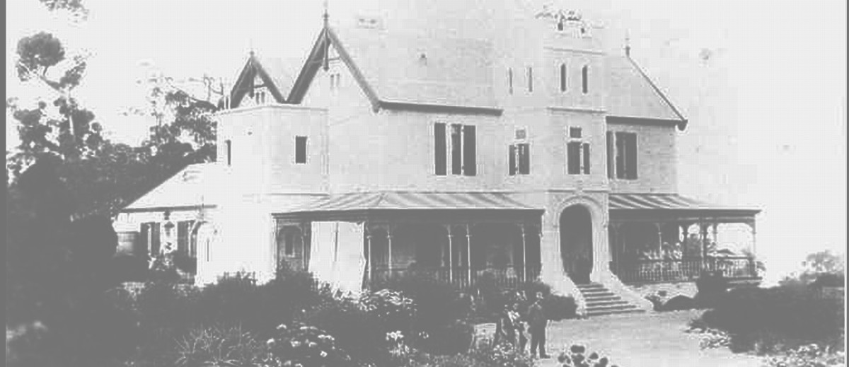 Image: A large, two-storey mansion stands at the top of a hill surrounded by trees and vegetation. A small group of men and women stand in front of the house