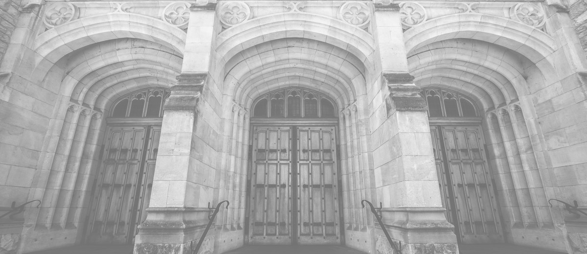 Image: The entrance to an ornate, historic building made of stone. Three large wooden doors are contained within corresponding stone archways