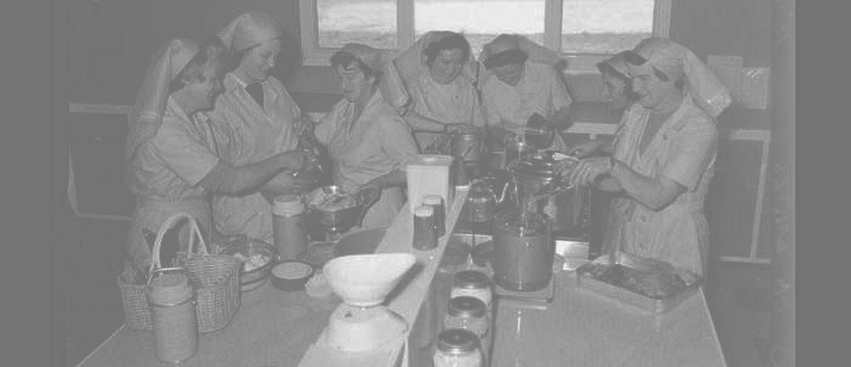 Image: A group of seven Caucasian women in 1960s attire that resemble nursing uniforms prepare and pack food in a small kitchen