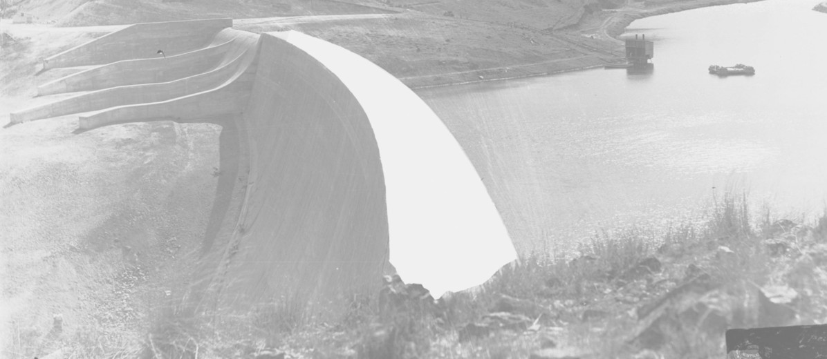Image: A large concrete dam located among rolling hills. To the right of the dam is a reservoir of water, and to its left a dry rocky chasm