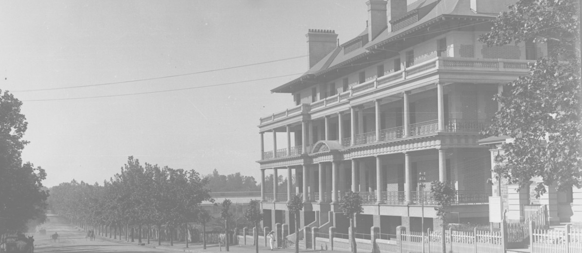 Image: A large, four-storey brick building sits alongside a dirt road lined with trees. Four horse-drawn carts are visible travelling along the street