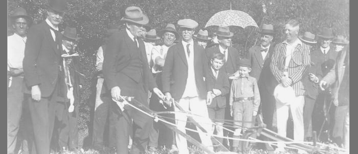 Image: A man in a hat and suit operates a plow in front of a small group of people