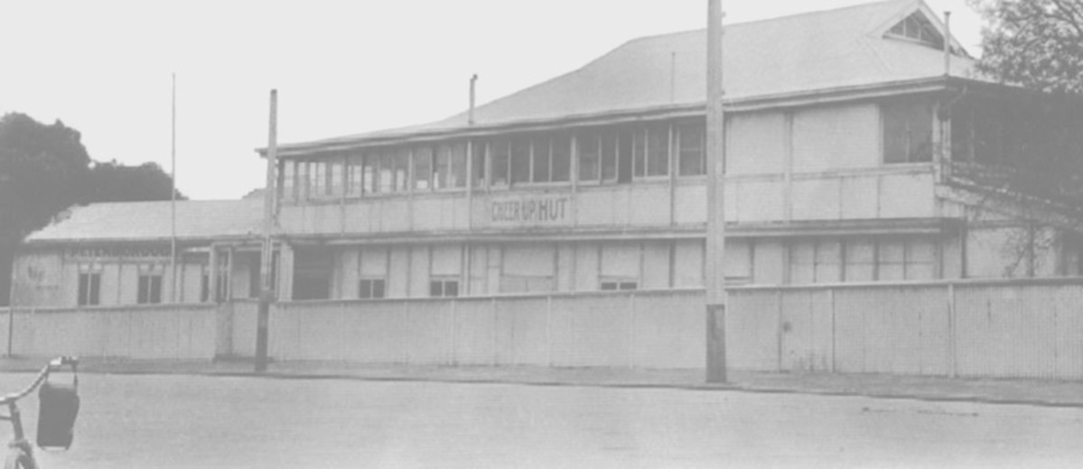 Image: Black and white photograph of a two storey building with a sign reading 'Cheer Up Hut' painted on the sign. A bag draped over the handlebars of a bicycle is just visible in the bottom left corner of the photograph. 