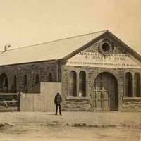 Image: a man in 1870s era clothing stands outside a small stone foundry with arched windows and doors and a medium pitched gable roof. To the left of the building is a fenced yard where more men work at various machinery next to a tall brick chimney.