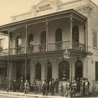 Image: a group of men, a woman and a boy in 1870s attire stand outside a two storey hotel with a balcony, arched windows, and a parapet sign which reads "A.KLAUER WHITE HEART HOTEL"