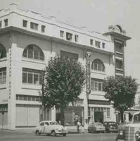 Image: a four storey white stucco building with large arched on its third floor. It sits on the corner of a tree lined street which is busy with 1950s era automobiles. 