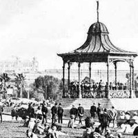 Image: A crowd of people in early twentieth century attire sit on the grass around a rotunda where a band plays