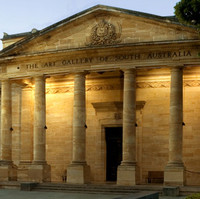 Image: The front of a large stone building with six columns flanking the entrance. A fountain and benches are arranged along stone sidewalks in front of the building