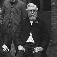 Image: Three elderly Caucasian men in late Victorian-era attire pose for a photograph outside a bluestone building. Two are seated while another stands. 