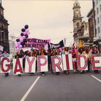 Image: men and women in 1970s era clothes march down a city street with pink and black balloons and signs reading "gay pride" and "homosexual liberation"